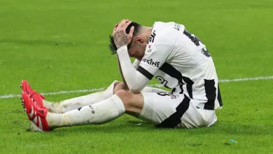 Beşiktaş forması giyen bir futbolcu, saha zemininde yere oturmuş şekilde başını iki eliyle tutuyor. Üzgün ve bitkin bir pozisyonda, gözleri yere eğik. Beyaz-siyah forma çamurlu, zeminde mücadele izleri dikkat çekiyor.